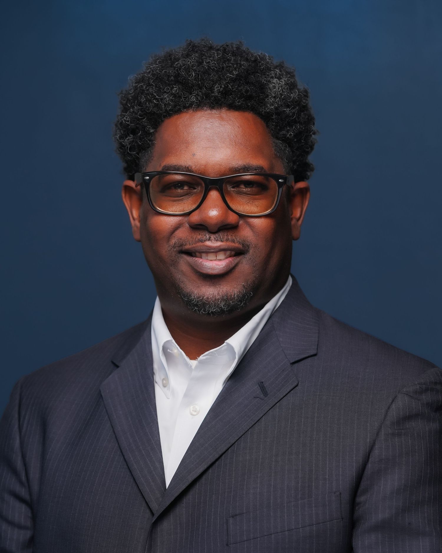 Professional headshot of a smiling man wearing glasses and a navy suit against a dark blue background.