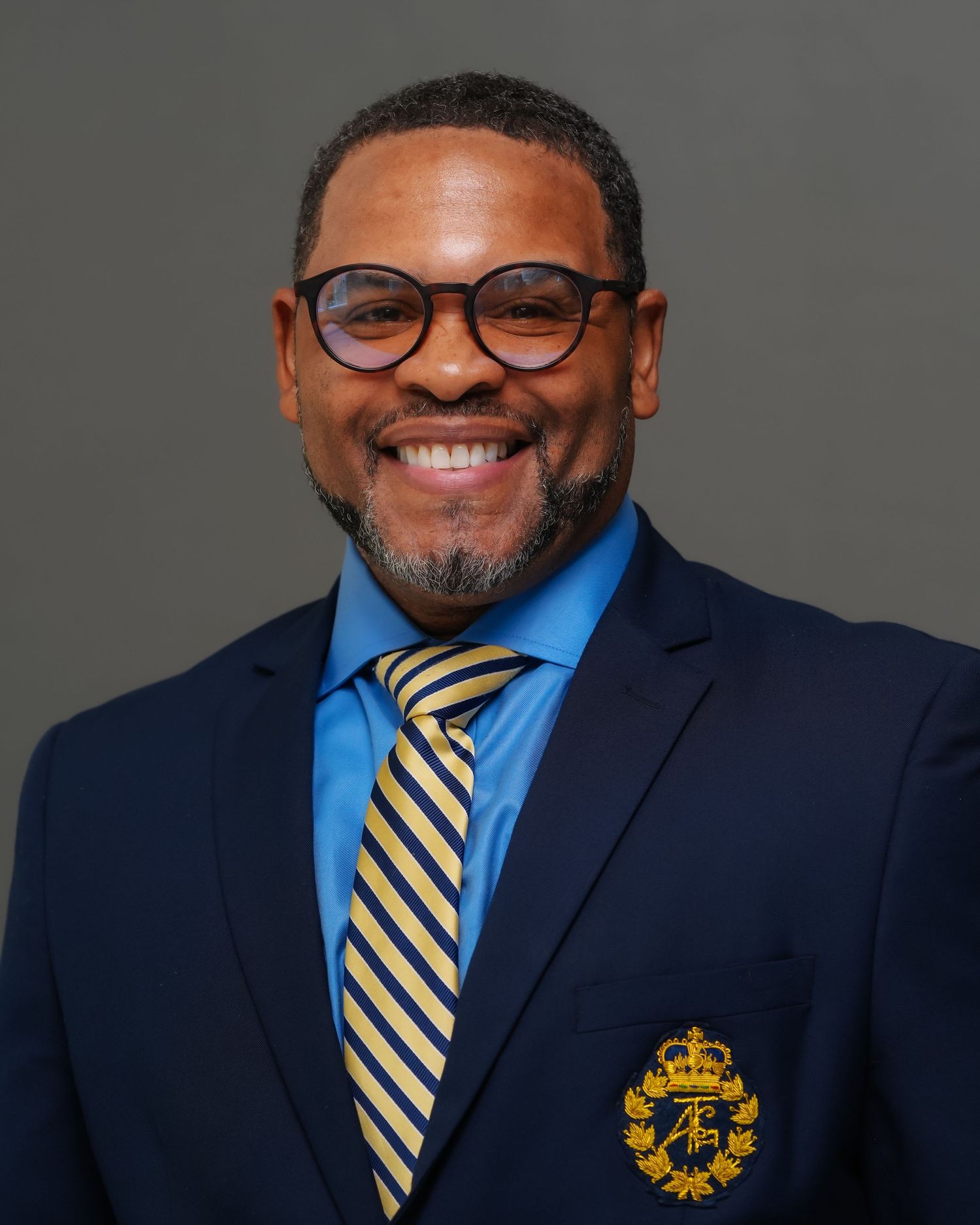 Professional headshot of a smiling man in a navy blazer with gold crest, blue shirt, and striped tie.