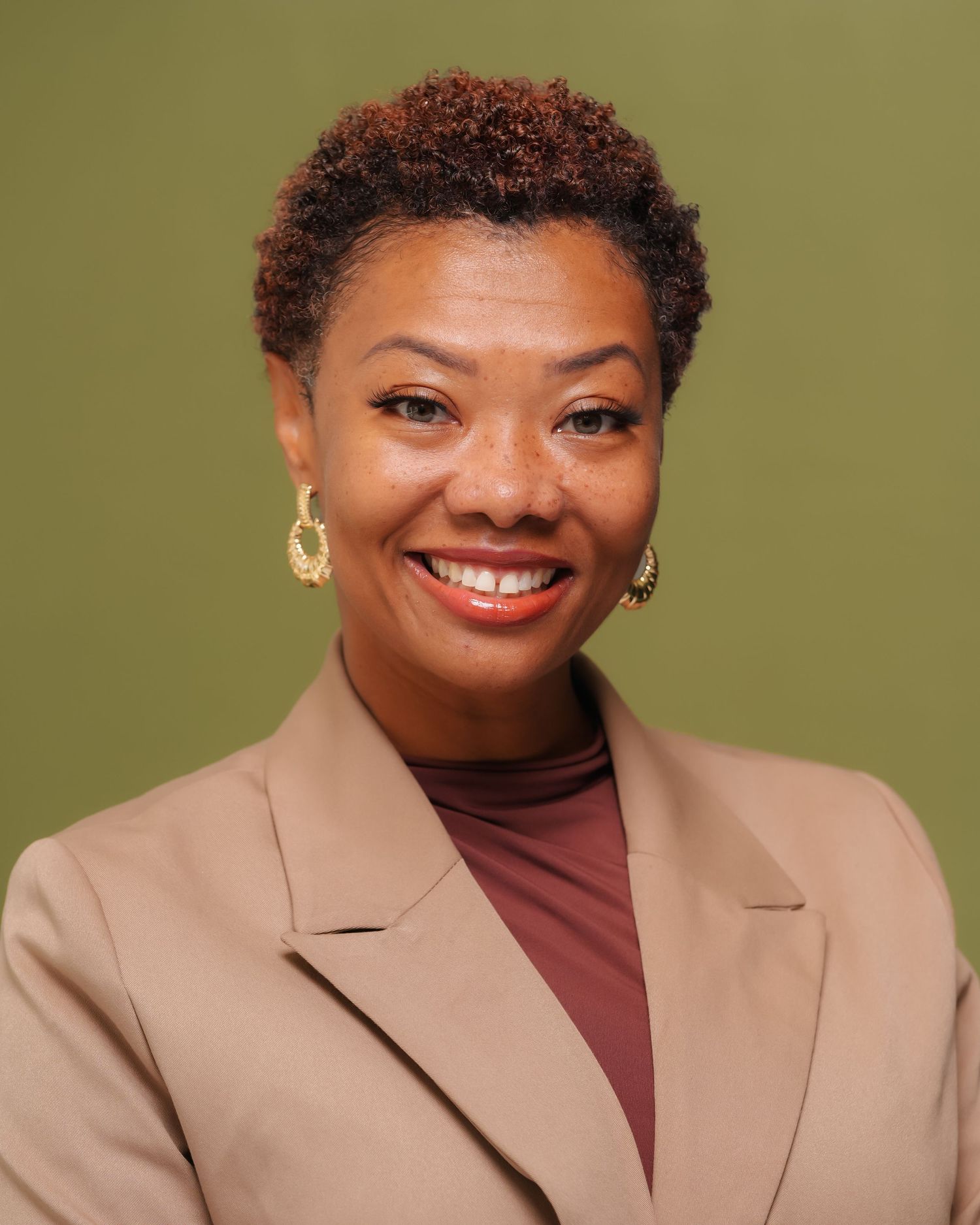 Professional headshot of a smiling woman in a beige blazer with gold earrings against a green background.
