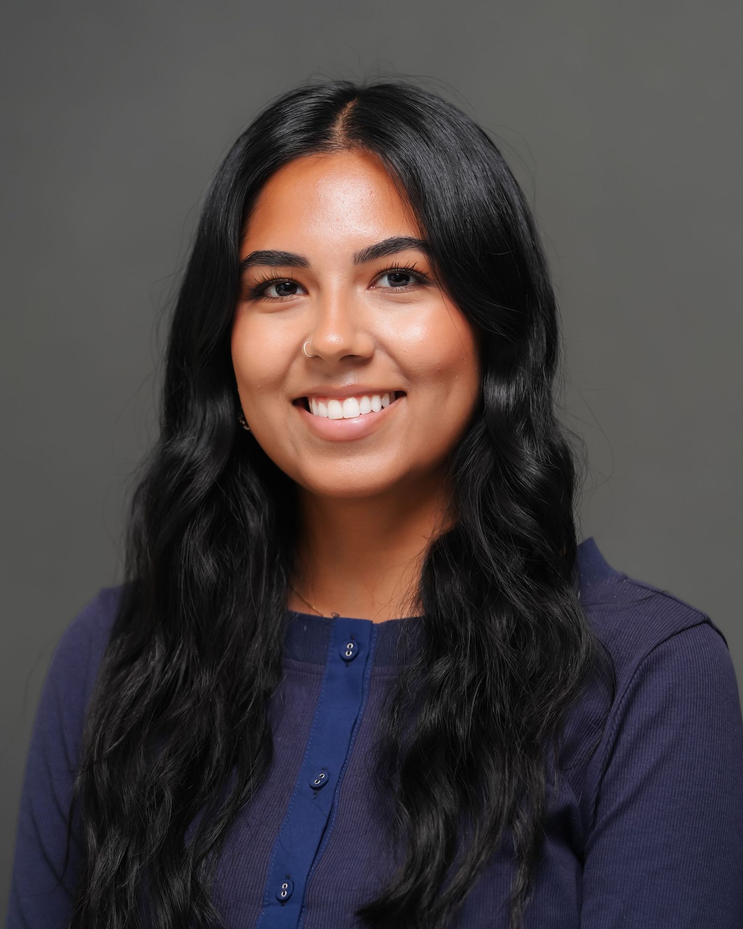 Young woman with long dark wavy hair smiling, wearing a navy blue top, professional headshot on gray background.