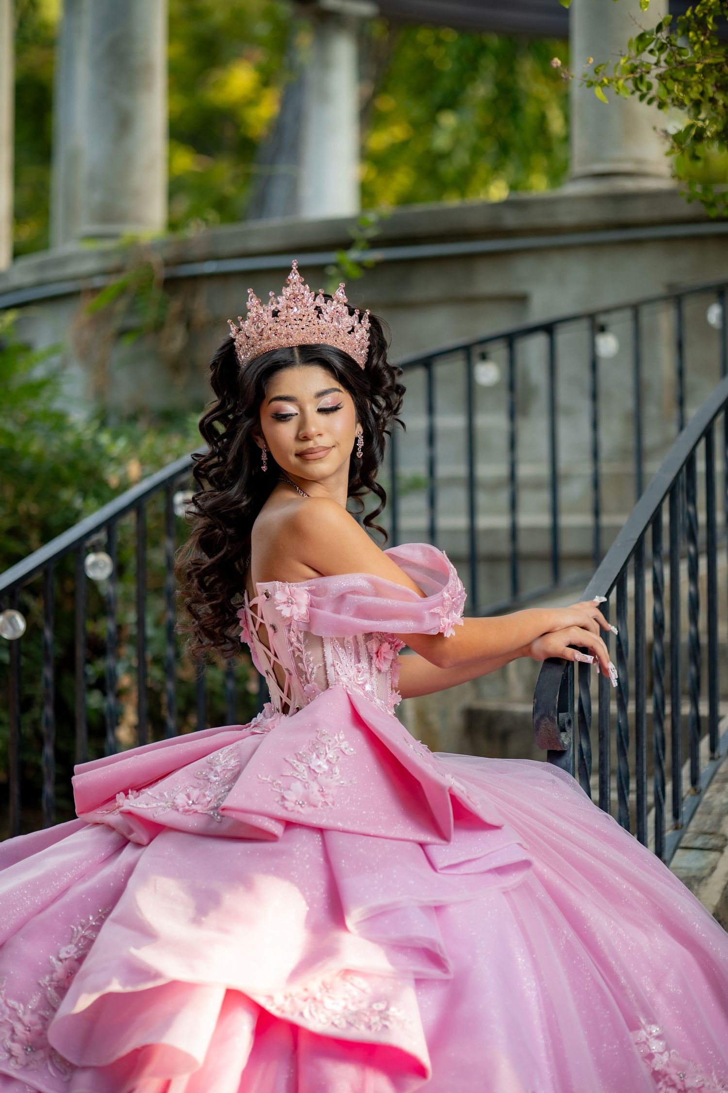 Beautiful young woman in pink quinceañera gown and crown poses on staircase in garden setting.