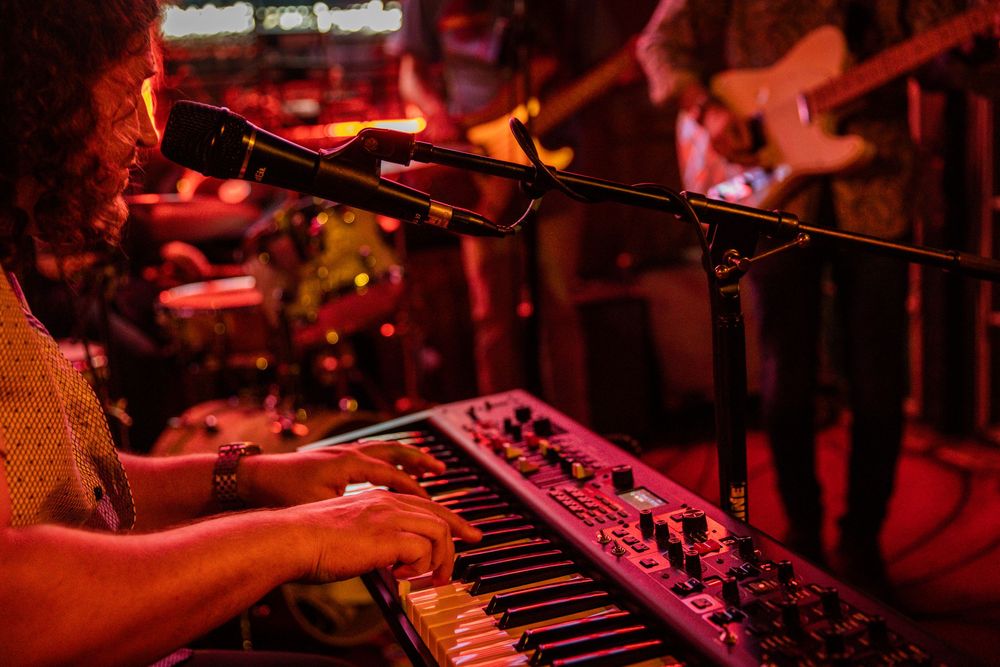 Musician plays a Nord keyboard in red stage lighting at a live music venue.