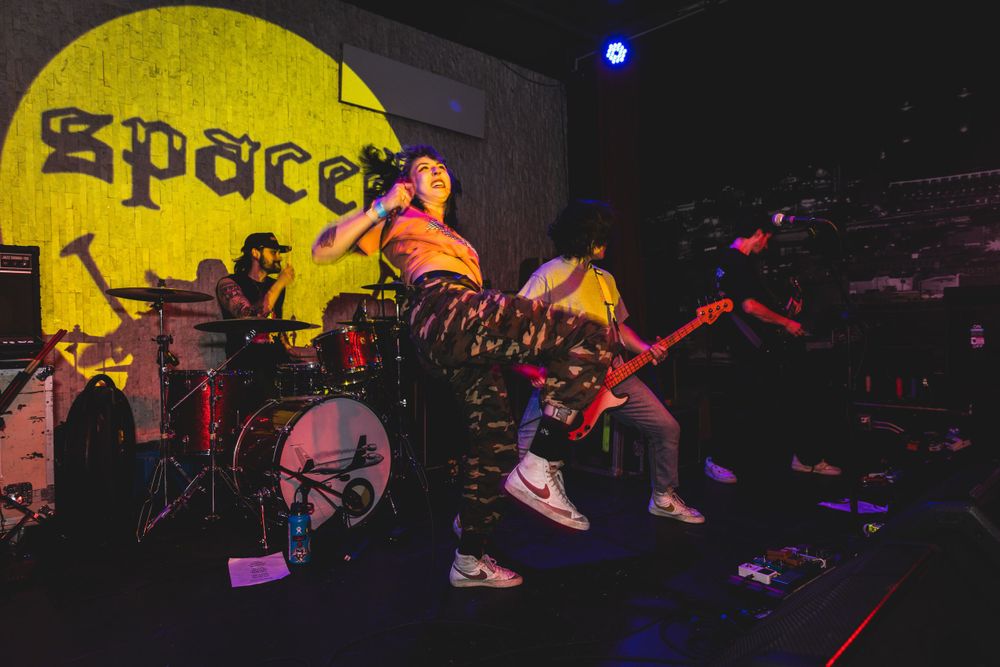 Rock band performs energetically on a dimly lit stage with colorful lighting effects and dramatic shadows.