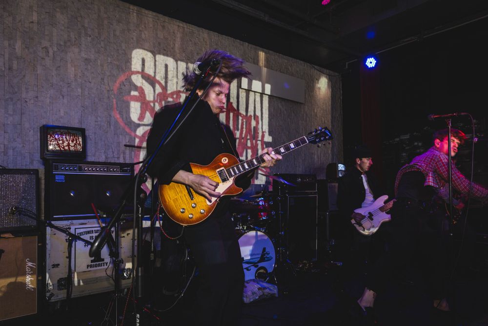 Musicians play instruments on a dark concert stage illuminated by blue and purple spotlights.