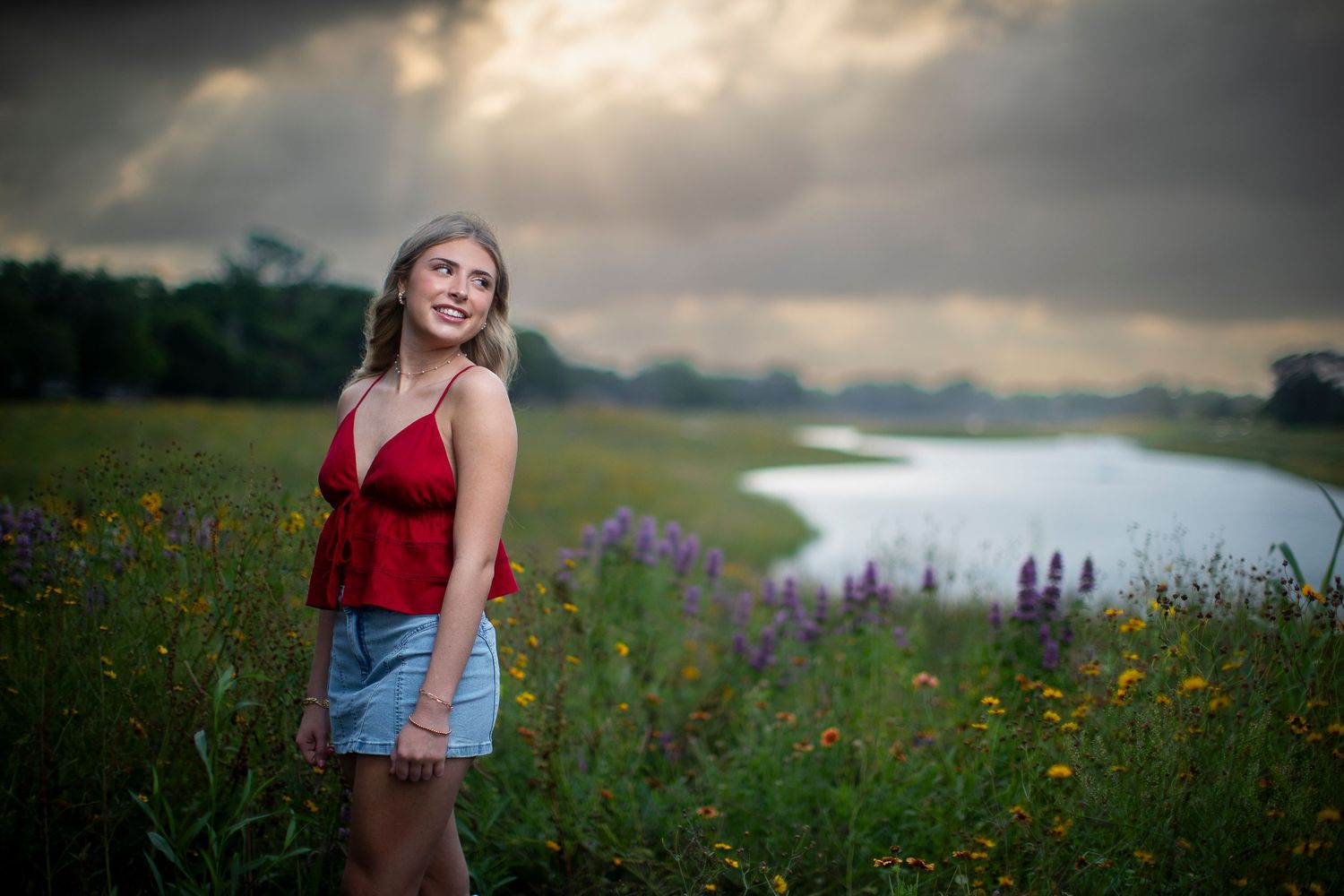 A person in a red dress poses in a wildflower meadow at sunset with dramatic cloudy skies in the background.
