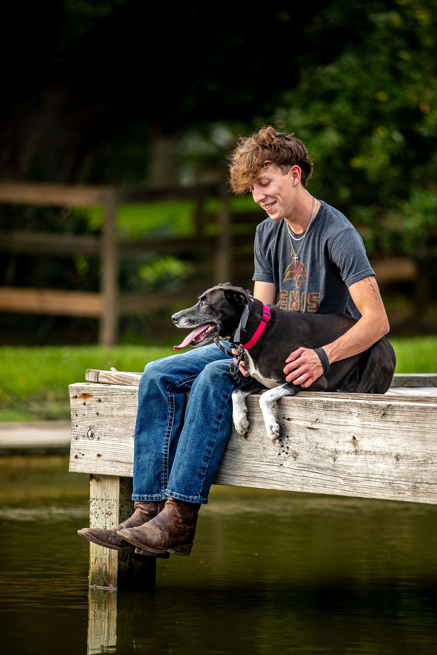 Person in jeans and t-shirt sits casually on a wooden dock by a lake on a sunny summer day.