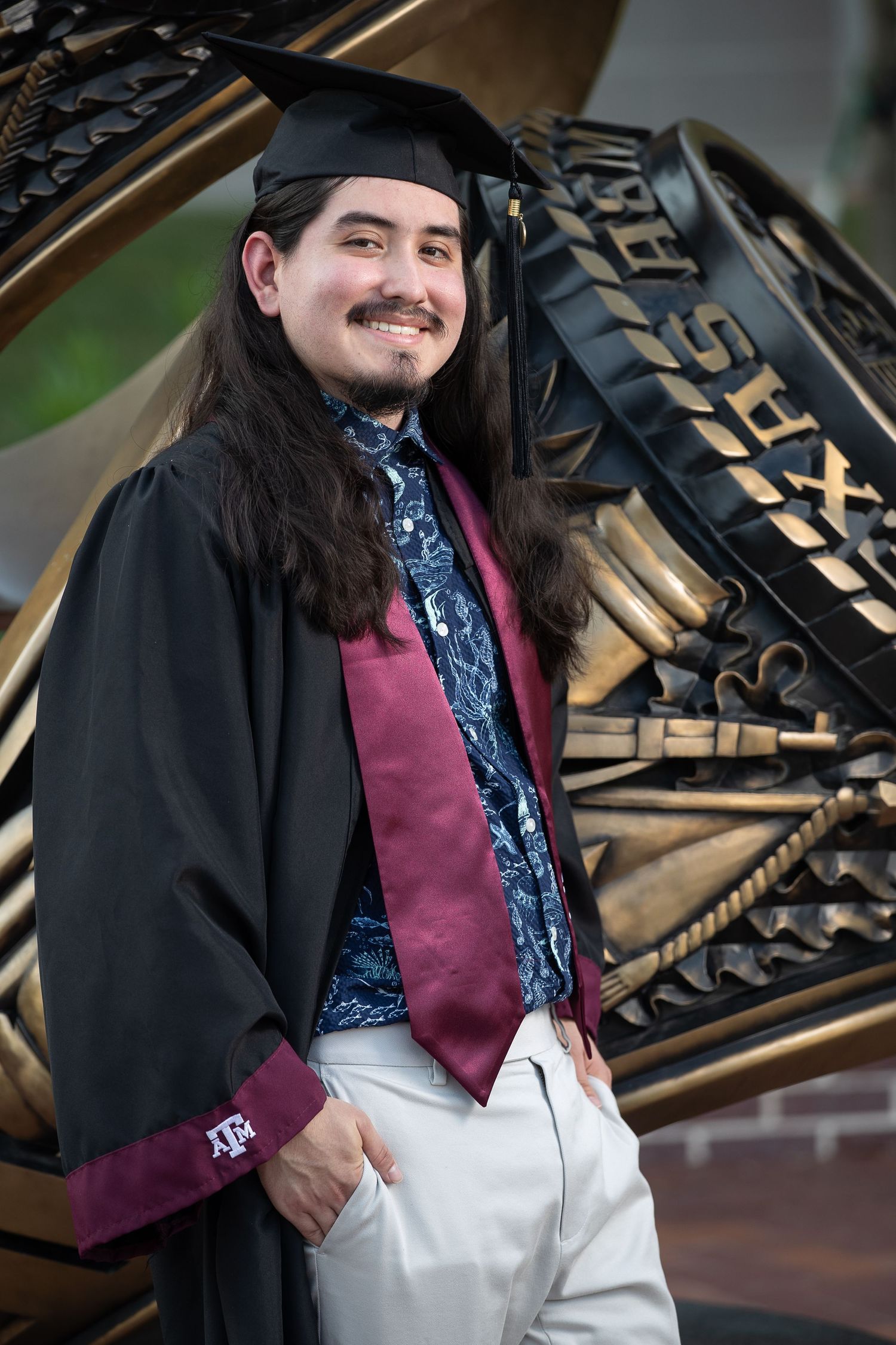 Graduate in cap and gown wearing burgundy stole and patterned shirt poses by ornate architectural detail.