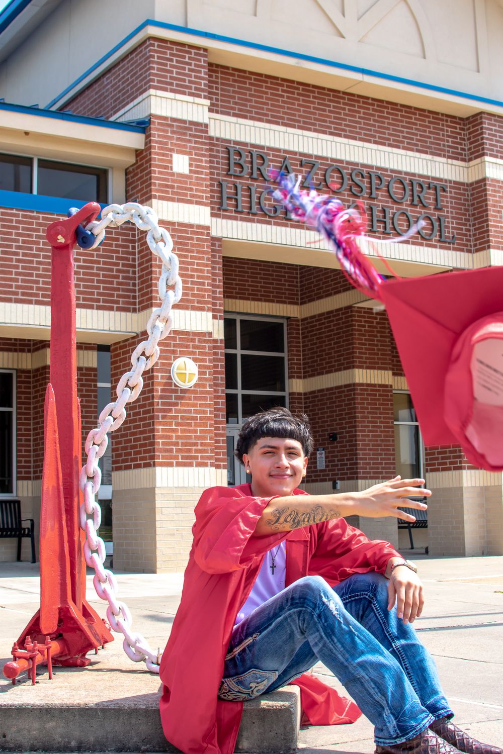 Person in red graduation gown sits casually on steps with decorated entrance of Brazosport High School behind them.