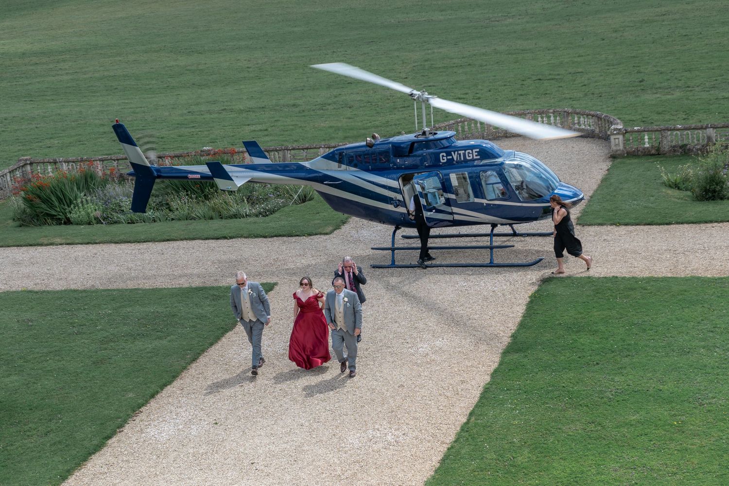 Blue helicopter landed on pathway with wedding party approaching in formal attire.