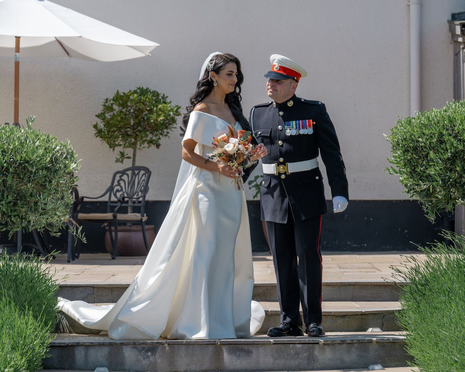 A military wedding couple poses elegantly on garden steps beneath a white umbrella.
