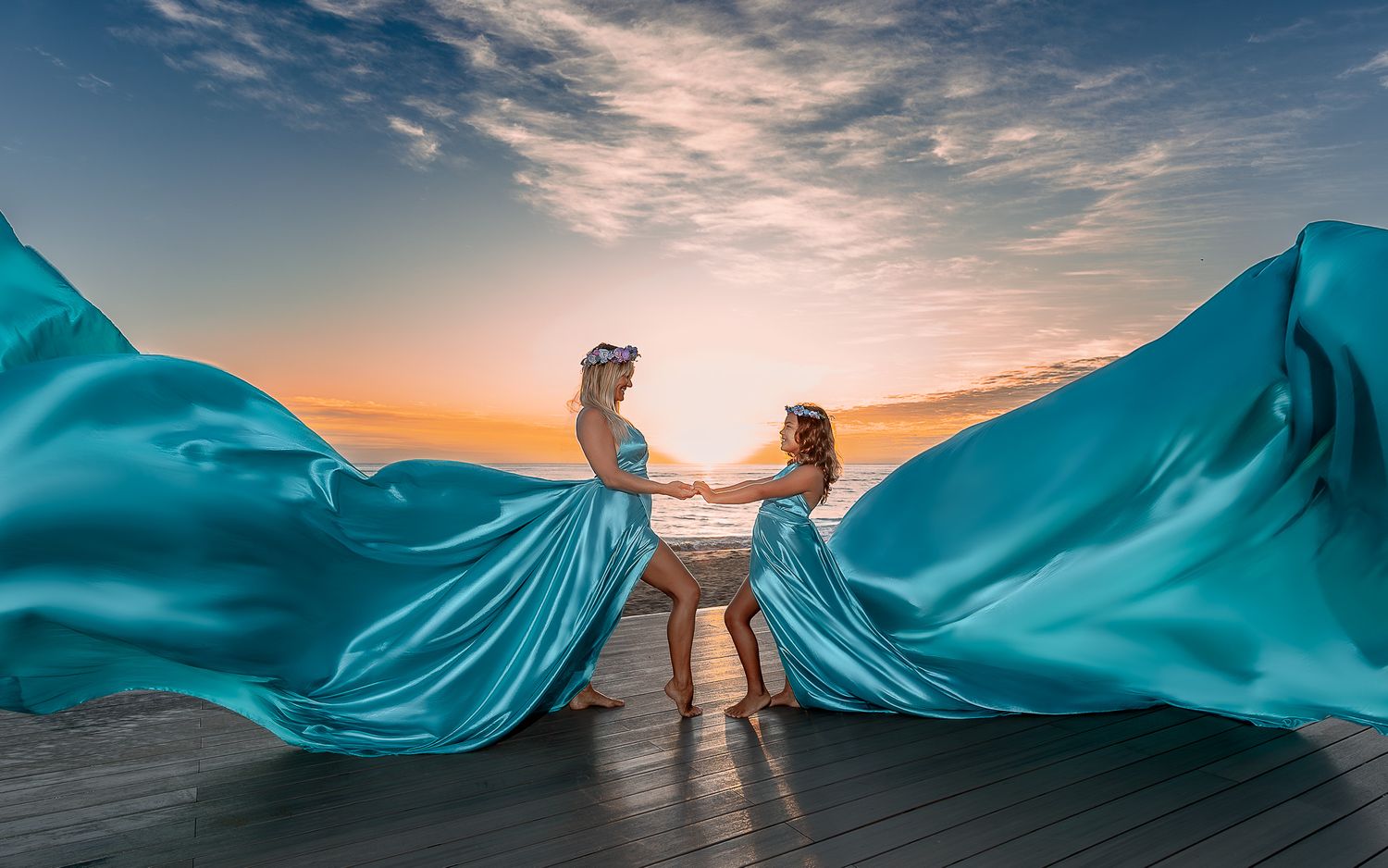 Two figures in flowing turquoise dresses pose dramatically against a sunset sky.