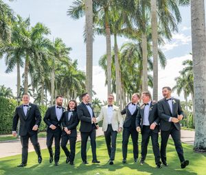 Groom in white tuxedo jacket poses with groomsmen in black tuxedos on tropical palm tree lined lawn.