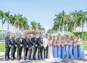 Wedding party poses in front of a fountain surrounded by palm trees, bridesmaids in blue gowns and groomsmen in gray suits.
