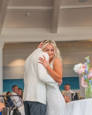 Bride in white gown shares emotional first dance embrace with groom in white suit at beachside reception.