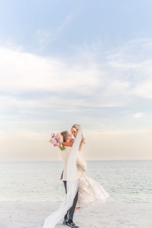 Groom lifts bride in wedding dress on beach at sunset, her veil flowing as she holds a pink bouquet.