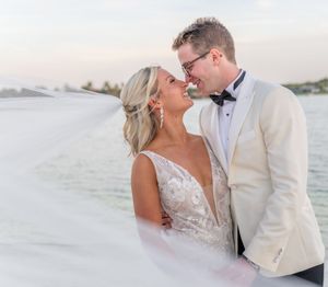 Bride and groom share a romantic moment smiling at each other outdoors with a flowing veil and waterfront backdrop.