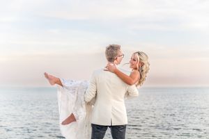 Groom in white suit carries bride in lace wedding dress on a serene beach at sunset.
