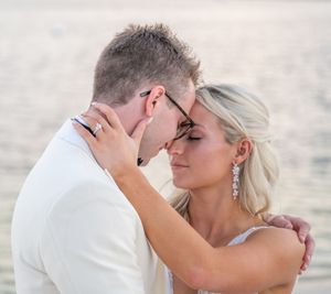 Bride and groom share intimate forehead touch near shimmering water at golden hour beach wedding.