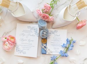 Elegant wedding stationery flat lay with pink and blue flowers, ring box, and white bridal heels on marble surface.