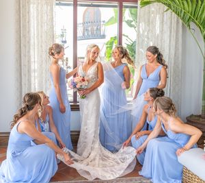 Bride in lace gown surrounded by bridesmaids in blue dresses adjusting her veil and train near a sunlit window.