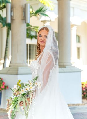 Beautiful bride in a white wedding gown and veil poses with tropical bouquet by elegant columns on her wedding day.