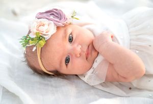 A baby in soft white clothing lies on a white blanket with delicate floral accents near their head.