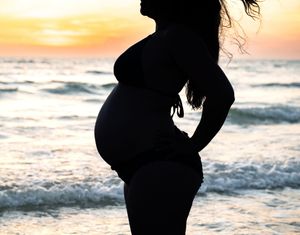 Silhouette of pregnant woman standing on beach against golden sunset sky and ocean waves.