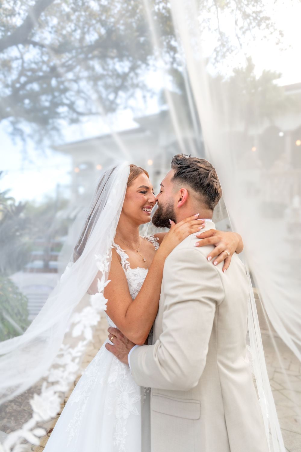 Newlywed couple sharing romantic kisses in a dreamy outdoor setting with flowing white veil and cream suit.