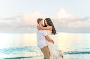 Romantic couple embraces at sunset on tropical beach with turquoise water and soft clouds in background.