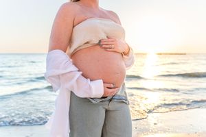 A pregnant belly silhouetted against a sunset beach backdrop with waves rolling in along the shore.