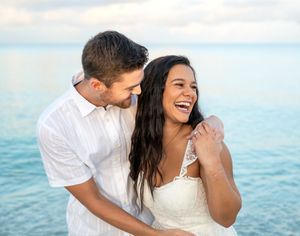 A joyful couple laughing together in a romantic moment by the ocean with blue water and sky in the background.