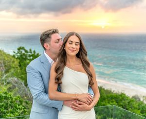 A romantic couple embraces on a cliff overlooking a beautiful turquoise ocean at sunset.