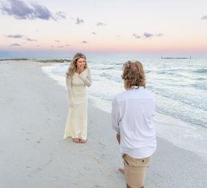 A couple shares a romantic moment on a sandy beach at sunset with turquoise waves and soft pink sky in the background.