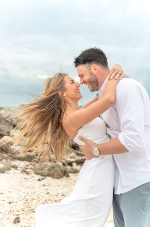 Romantic couple in white outfits embracing and laughing together on a rocky beach with wind-swept hair.