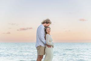 A couple embraces on the beach at sunset with the ocean waves and pastel sky in the background.