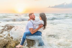 Couple embraces on beach rocks during golden sunset as ocean waves crash around them on a romantic evening.