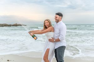 A couple celebrating on the beach at sunset while popping open a bottle of champagne.