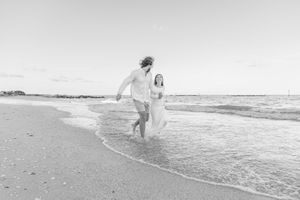 A couple in white clothing runs joyfully along the beach shoreline in a dreamy black and white photograph.