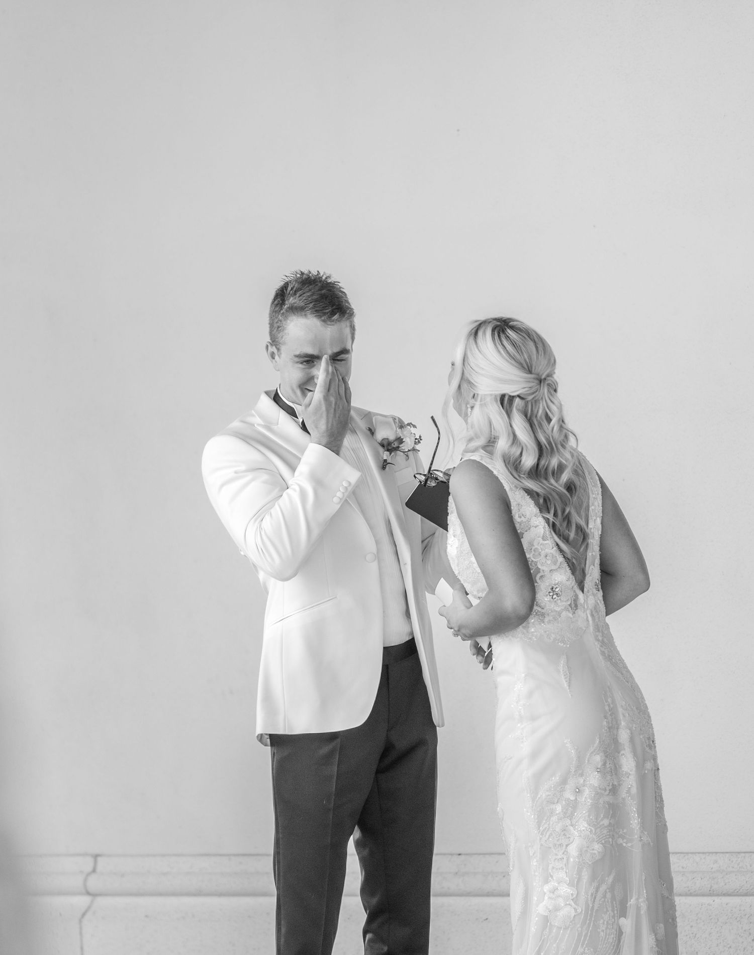 Groom covers his mouth in emotional reaction seeing bride for first time in black and white wedding photo.
