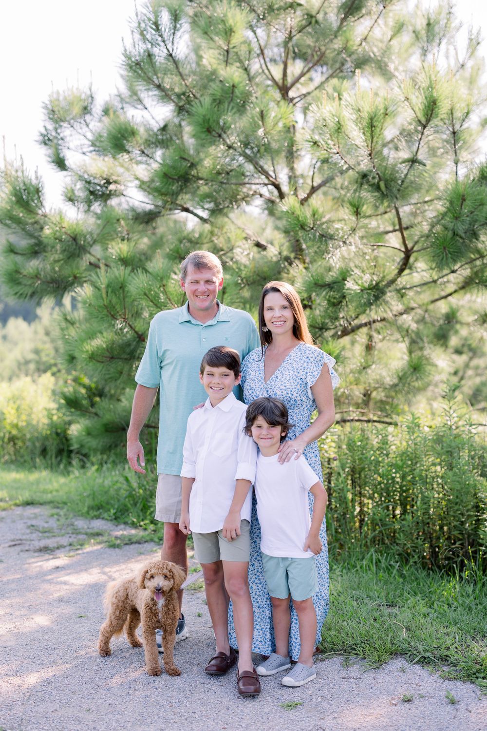 A family of three poses with their brown goldendoodle dog on a nature trail during a summer photo session.
