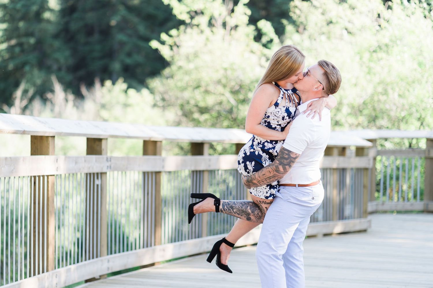 Two people share a romantic embrace on a wooden footbridge on a sunny day in nature.