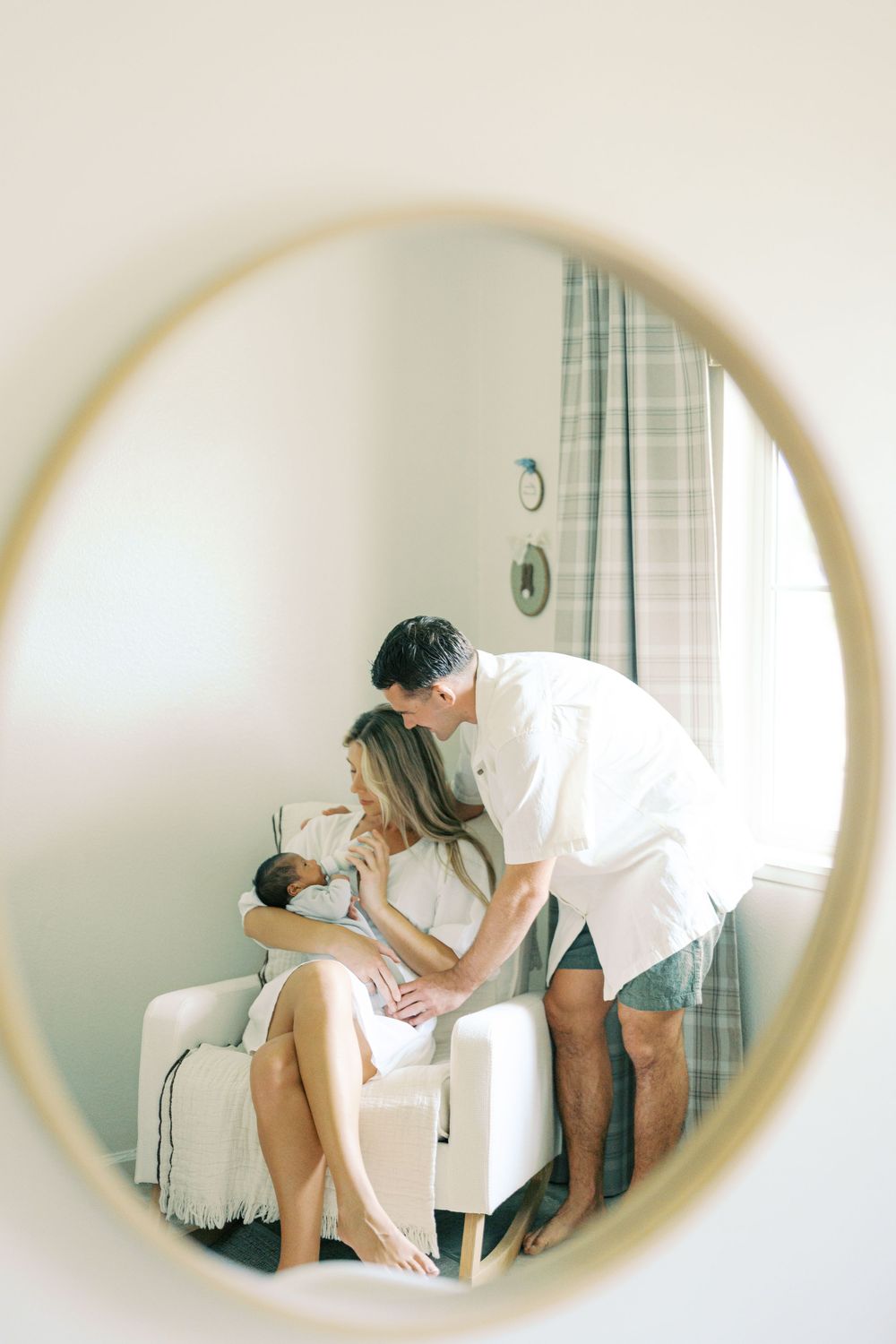 A couple admires their newborn baby, reflected in a round mirror, in a softly lit nursery room.