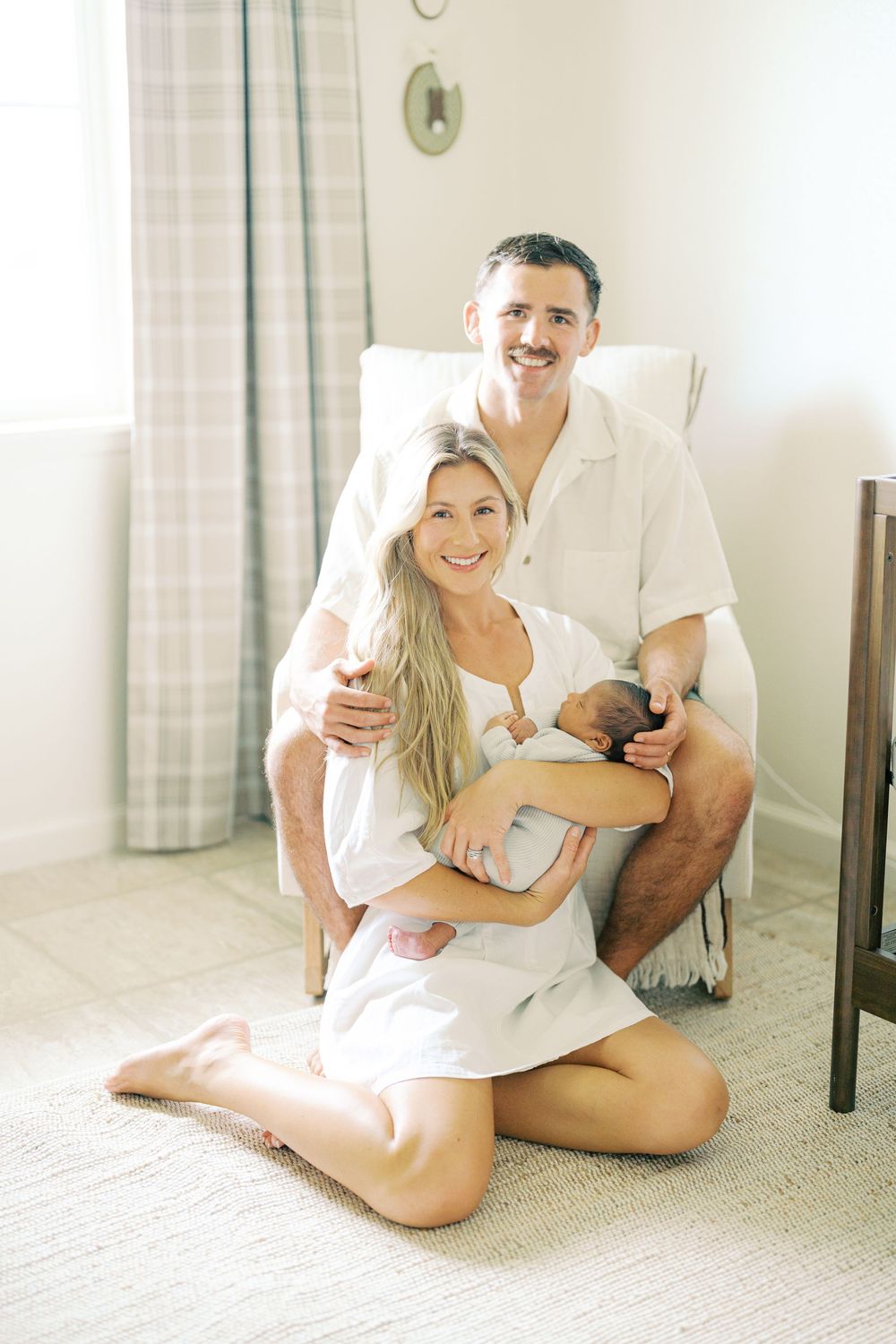 Happy couple sitting on floor holding newborn baby in bright nursery room, smiling at camera.