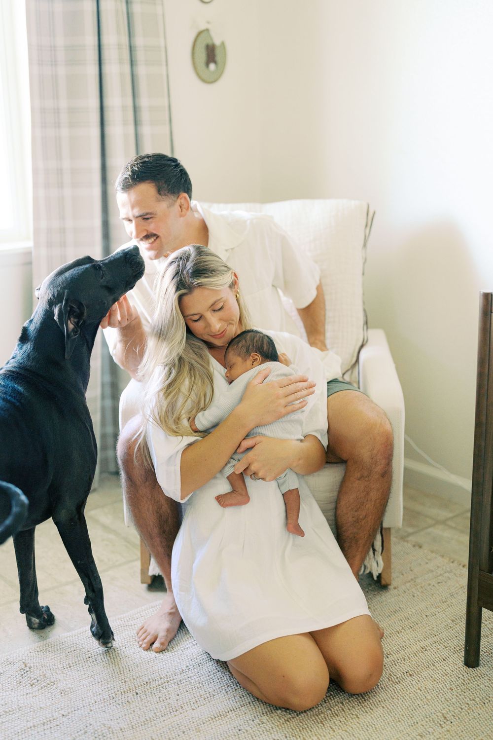 A happy family with a newborn baby and black dog sitting together in a cozy nursery chair at home.