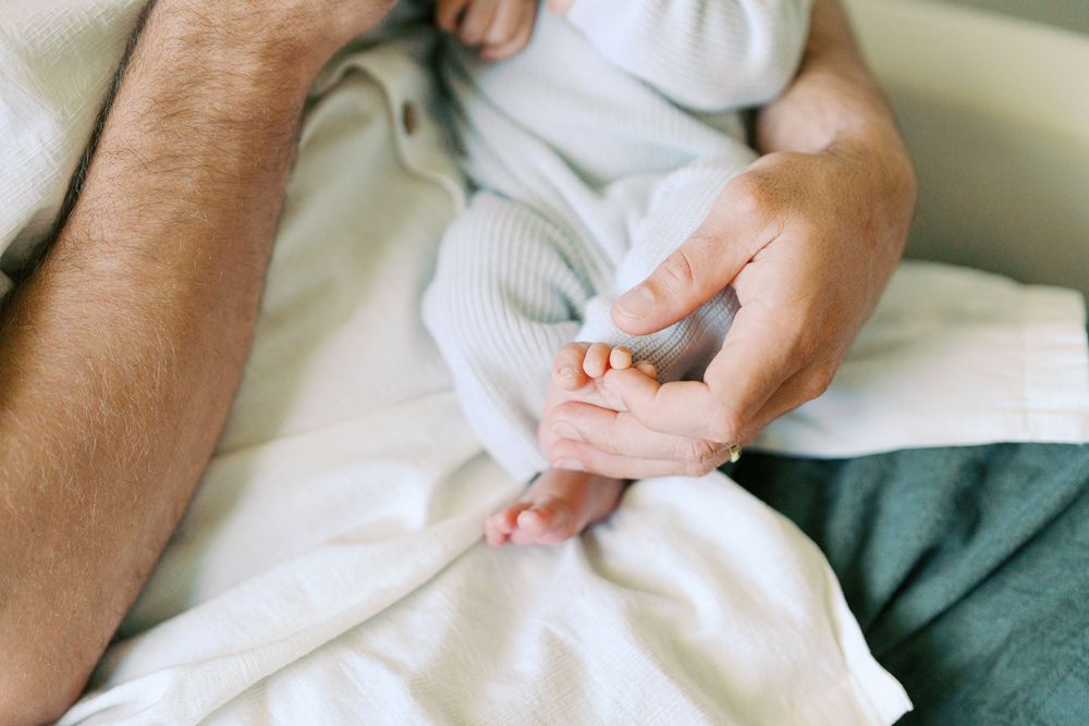 Adult hand gently holding tiny newborn baby feet on a soft white blanket in a tender parenting moment.