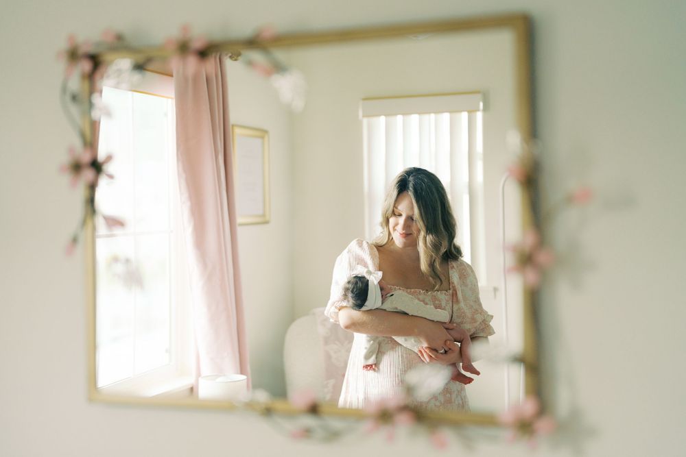 A bride in a white dress holds her newborn baby, reflected in a floral-framed mirror in a softly lit room.