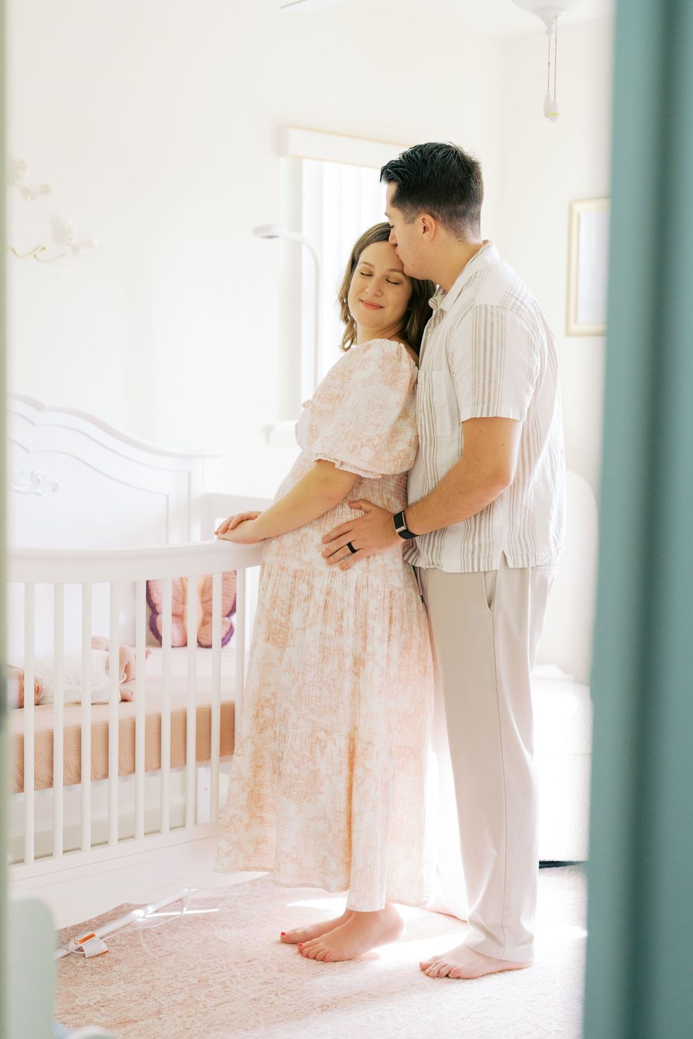 Couple standing together by a white crib in a softly lit nursery, woman in floral dress, man embracing her from behind.