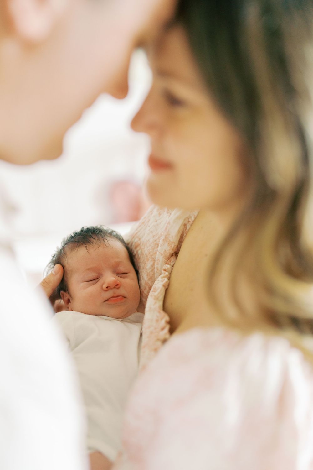 Two parents lovingly gazing at their sleeping newborn baby wrapped in white, in a soft, warm light.