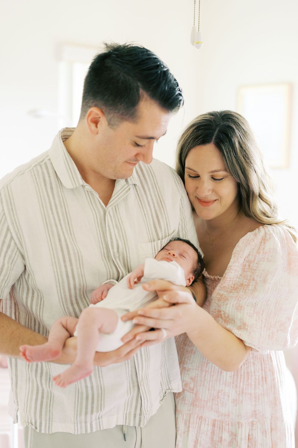 New parents lovingly cradle their sleeping newborn baby, sharing a tender family moment in a bright, airy room.