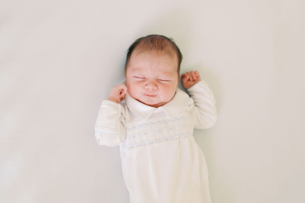 Newborn baby in white outfit lying on white blanket, eyes closed, fists raised in a stretching pose.